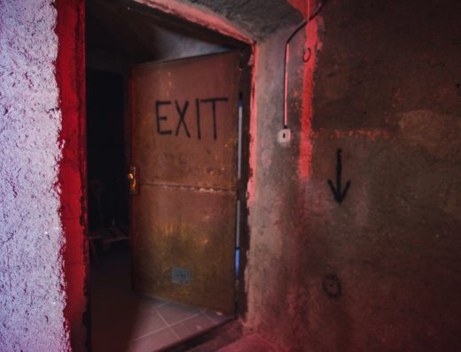 Porte métallique entrouverte avec l’inscription « EXIT » dans un couloir sombre éclairé en rouge.